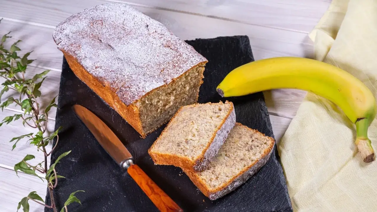 Saftiger Bananen Buttermilch Kuchen, bestäubt mit Puderzucker, neben einer reifen Banane und einem Messer.