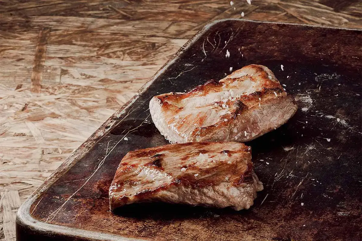 Zwei saftige Duroc Secreto-Steaks, perfekt gegrillt und mit grobem Salz bestreut, liegen auf einem rustikalen Blech. Die Zubereitung verspricht ein Genusserlebnis.