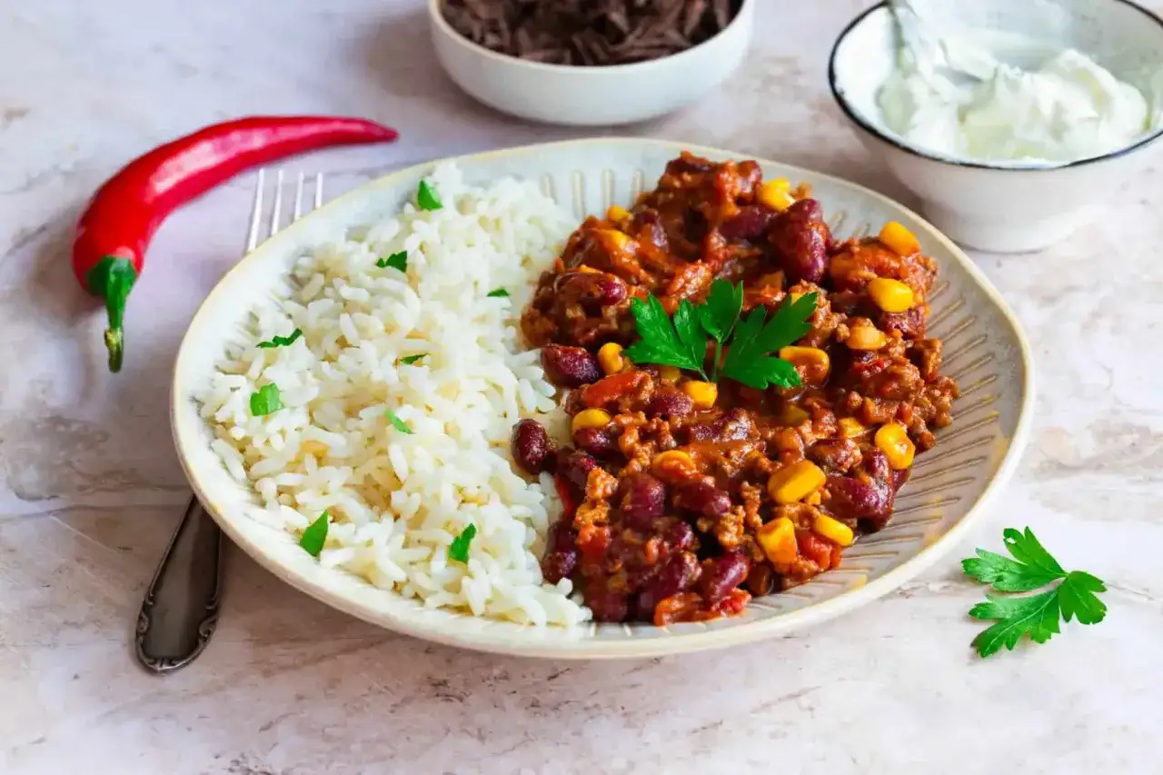 Herzhaftes Chili con Carne mit Reis als Beilage, garniert mit Petersilie. Daneben ein roter Chili und ein Schälchen mit Sauerrahm.