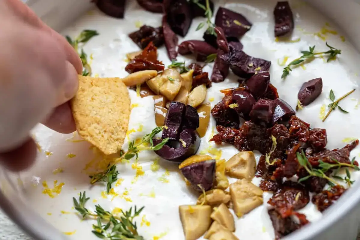 Ein Keks wird in einen cremigen Tomaten Feta Dip getunkt, garniert mit Oliven, getrockneten Tomaten und Thymian.