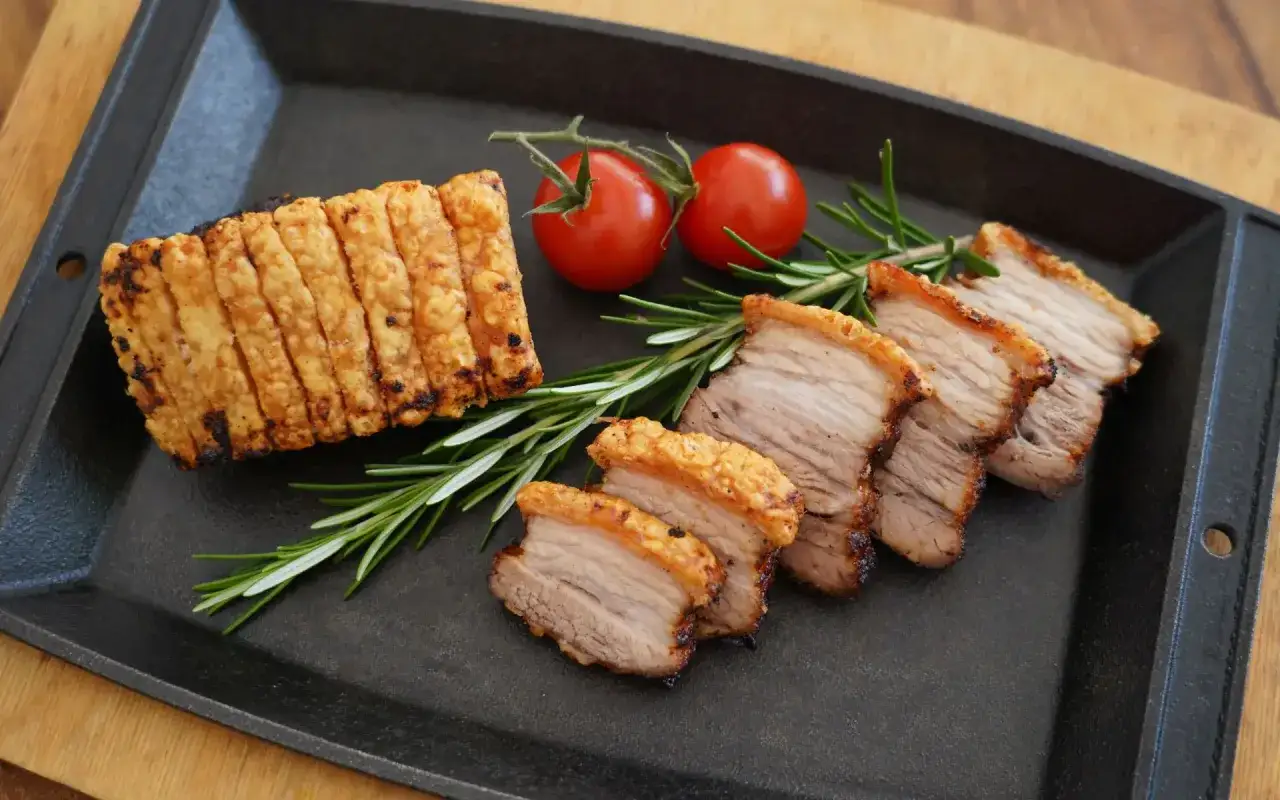 Knuspriger bauchspeck im airfryer, perfekt gegart mit krosser Schwarte. Daneben liegen saftige Scheiben, garniert mit Rosmarin und Kirschtomaten.