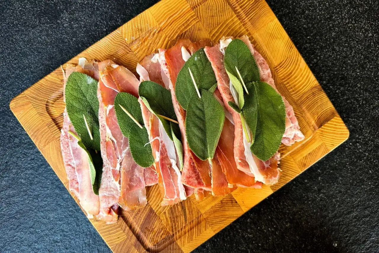 Zutaten für Saltimbocca alla Romana Rezepte: dünne Scheiben Kalbfleisch, umwickelt mit Prosciutto und Salbeiblättern, auf einem Holzbrett.
