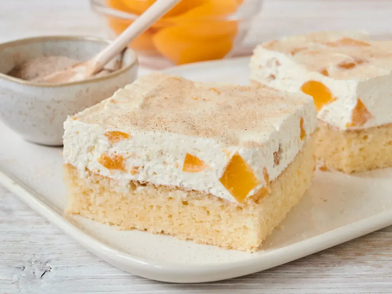 Ein St&uuml;ck saftiger Pfirsich Schmand Kuchen mit Zimt bestreut. Daneben eine Schale mit Pfirsichst&uuml;cken und Zimt.