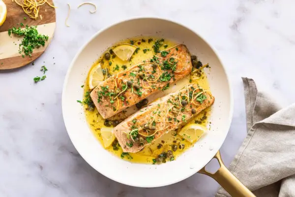 Zwei saftige Lachsfilets in einer Pfanne mit Zitronenscheiben, Kapern und Kräutern. Ein köstlicher Lachs mit Mangold-Aroma.