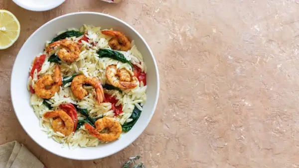 Ein Teller mit Orzo-Nudeln, Garnelen, Spinat und Tomaten, garniert mit Pfeffer. Dazu eine Zitronenspalte.